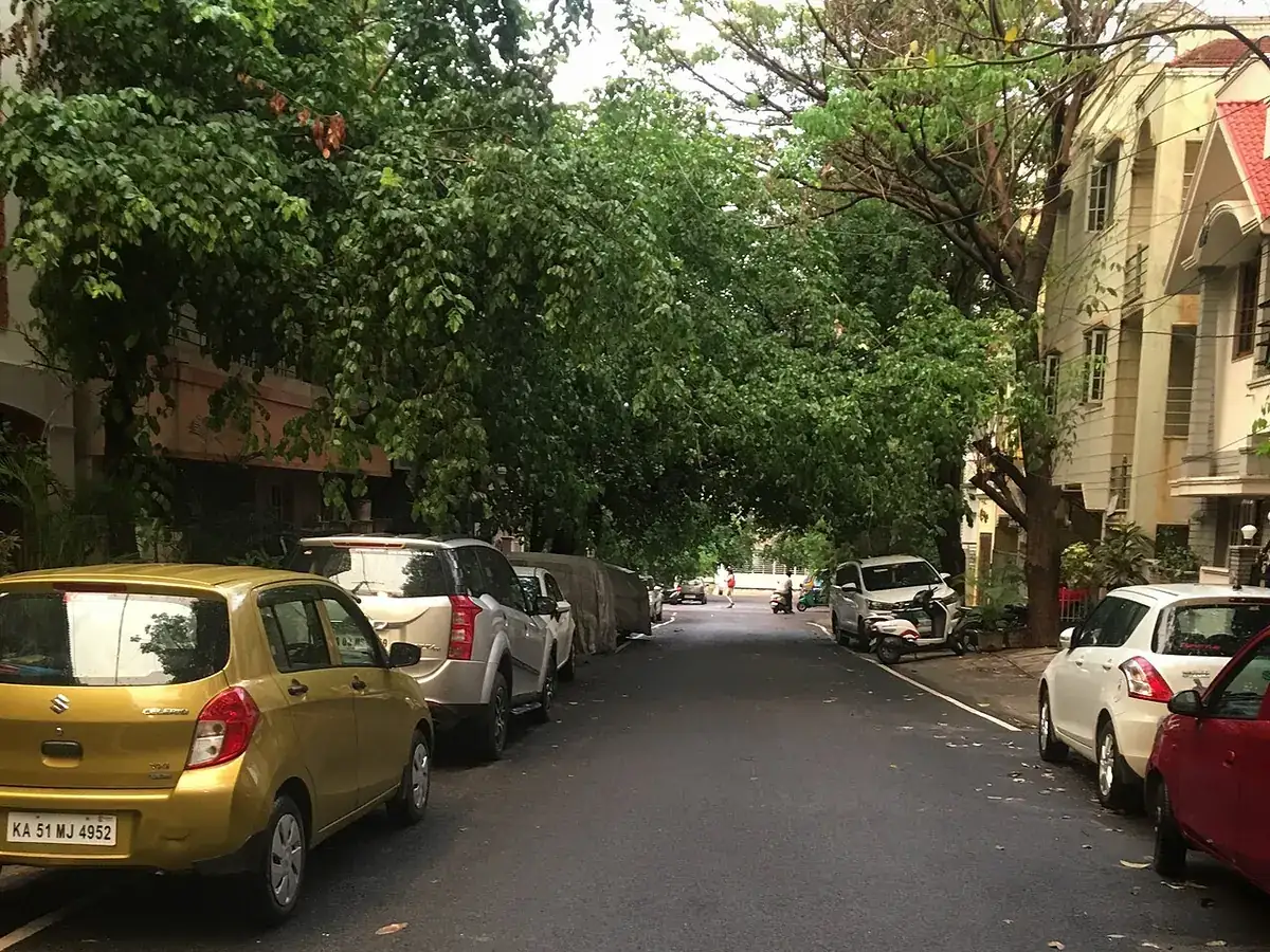 A quiet, tree-lined residential street in HSR Layout Sector 3, with cars parked along both sides and houses behind the trees