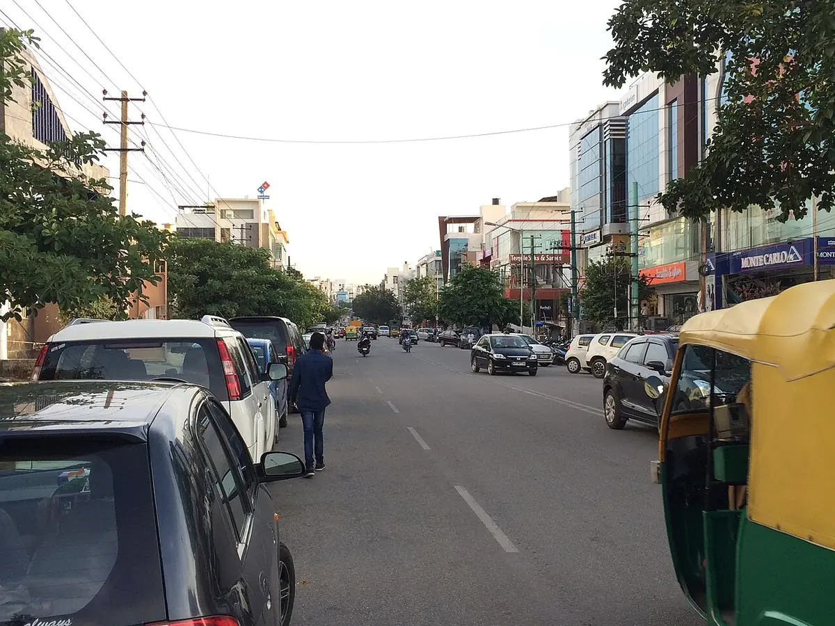 27th Main Road in HSR Layout Sector 1: a wide road lined with shops and multi-storey commercial buildings, with cars and an auto-rickshaw in the foreground